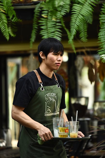 A young man with short black hair, wearing a black T-shirt and a green apron featuring a botanical logo, is holding a tray with several glasses containing beverages and straws. He is standing outdoors in a garden or patio setting, surrounded by lush, hanging fern plants with bright green, textured fronds overhead. In the background, there are blurred potted plants, wooden furniture, and natural light filtering through the greenery. The scene suggests a relaxed outdoor environment, with the individual engaged in serving drinks, indicating garden maintenance or outdoor hospitality work, consistent with gardening or landscaping themes. Gardeners Battersea is mentioned as part of the context for outdoor care and garden services.